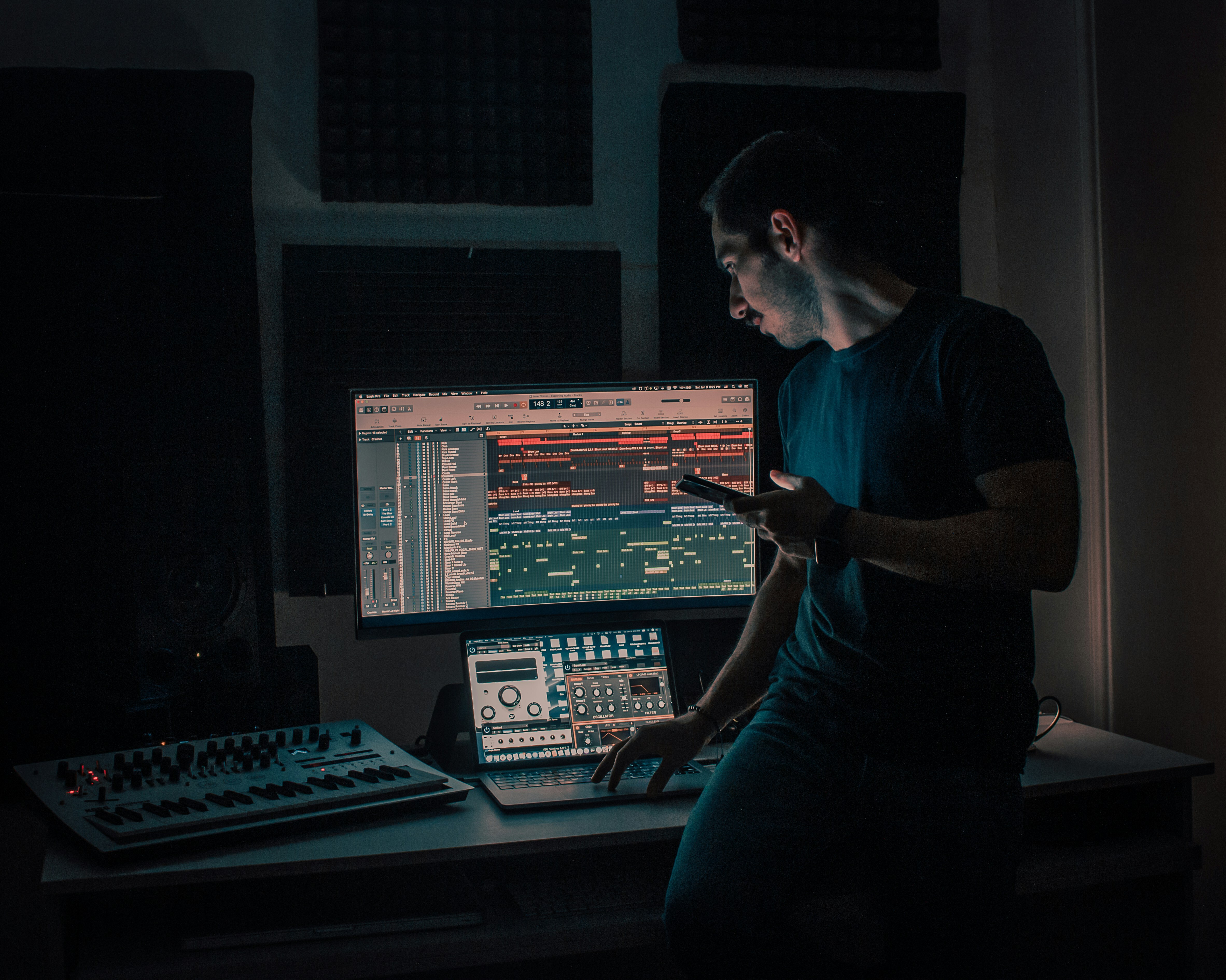 Fot. Rezli. Mężczyzna w studiu muzycznym stojący przy biurku z kontrolerem MIDI, laptopem i dużym monitorem wyświetlającym ścieżki dźwiękowe, trzymający w dłoni smartfon.