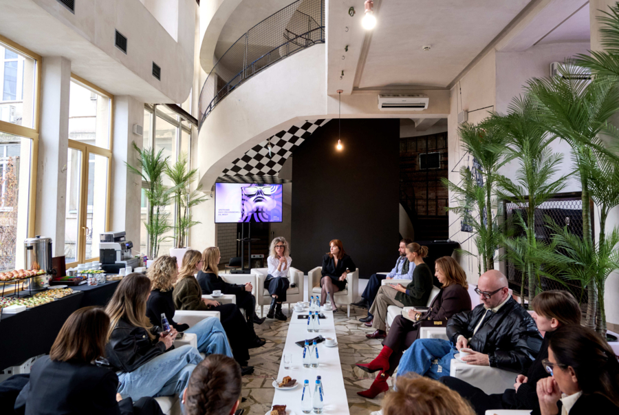 Grupa ludzi siedzi w fotelach wokół długiego, niskiego stolika podczas panelu dyskusyjnego w jasnym, architektonicznym wnętrzu. Po lewej stronie przy dużych oknach znajduje się stół z cateringiem i napojami, po prawej stoją wysokie palmy w donicach, a w tle widać kręcone schody oraz ekran z wyświetlaną prezentacją.