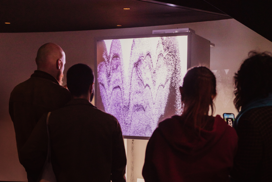 Scena galerii sztuki, widok od tyłu na cztery osoby obserwujące dużą, pionową instalację wideo w ciemnym pomieszczeniu. Ekran wyświetla jasne, abstrakcyjne, fakturowane wzory przypominające kaskady ziarnistego pigmentu w odcieniach fioletu i bieli. Mały rzutnik jest widoczny nad ekranem. Po prawej stronie jedna z osób trzyma smartfon, nagrywając wystawę. Widzowie stoją przed ekranem, oświetleni głównie jego blaskiem.