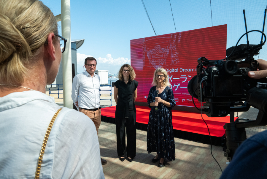 An outdoor event on a red podium on a sunny day. Three people—a man in a white shirt, a woman in a black jumpsuit, and a woman in a floral dress holding a microphone—are standing in front of a large red LED screen displaying Japanese and English text ("Digital Dreamers"). A port and the sea are visible in the background. In the foreground, the blurred back of a woman and a television camera can be seen.