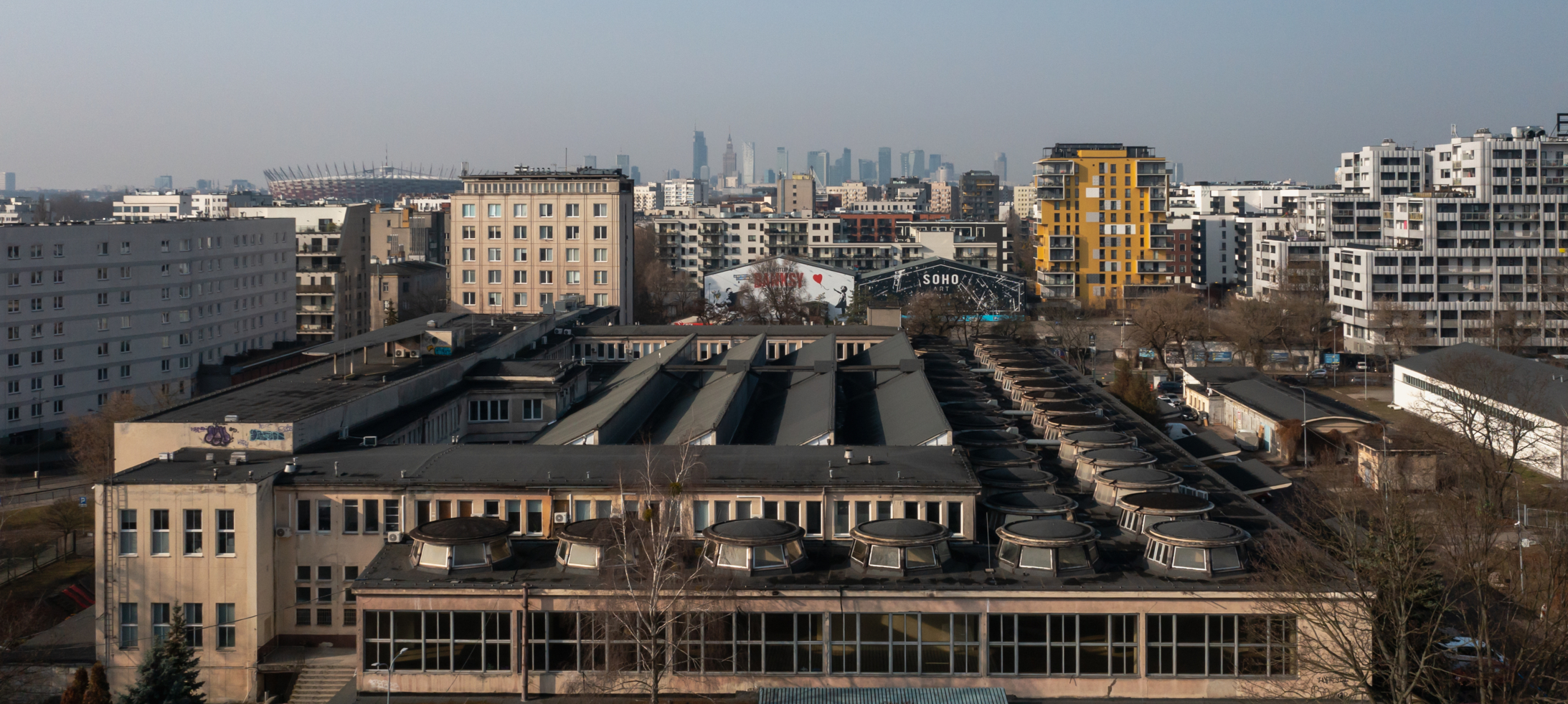 Widok z góry na niski, rozległy budynek z okrągłymi świetlikami na dachu w miejskiej otulinie. W tle, na horyzoncie, rozpościera się panorama miasta z wieżowcami i widoczną po lewej stronie sylwetką Stadionu Narodowego, a wokół budynku znajdują się osiedla mieszkaniowe oraz pojedyncze bezlistne drzewa.