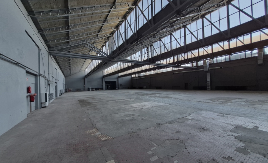 Wnętrze dużej, pustej hali przemysłowej z wysokim stalowym dachem i dużymi oknami. Wzdłuż ścian widoczne są drzwi, instalacje techniczne i gaśnice, a na posadzce zachowały się fragmenty starej, zniszczonej, płytkowej podłogi.