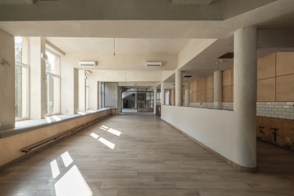 Wnętrze pustego, nowoczesnego lokalu w stylu industrialnym z drewnianą podłogą. Po lewej stronie rząd dużych okien, przez które wpada jasne słońce, rzucając cienie na podłogę, oraz długie, szare siedzisko okienne. Po prawej widać betonowe filary, białą ladę oraz ścianę wykończoną jasnym drewnem i białymi kafelkami. W głębi przeszklona ściana z wejściem. Surowy sufit z żarówkami i klimatyzatorami.