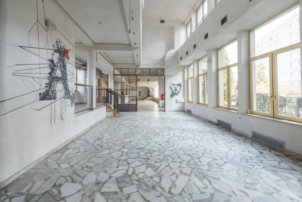 Jasne, puste wnętrze z posadzką z mozaikowego lastryko. Po lewej stronie na białej ścianie znajduje się duży, geometryczny czarno-czerwony mural przedstawiający abstrakcyjną konstrukcję. Po prawej ciągnie się rząd wysokich okien w drewnianych ramach, przez które wpada naturalne światło. W głębi widoczna jest przeszklona ścianka działowa z drzwiami oraz niewielki graficzny portret na ścianie. Przestrzeń jest surowa, jasna i przestronna.