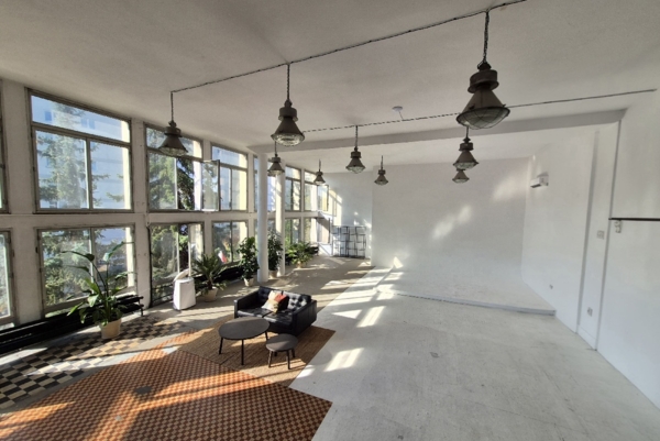 Jasne studio fotograficzne z białą cykloramą po prawej stronie. Po lewej ciągnie się ściana dużych okien wpuszczających naturalne światło słoneczne. Na mozaikowej podłodze ustawiono strefę wypoczynkową z czarną skórzaną sofą, okrągłymi stolikami i dywanem. Wzdłuż okien stoją zielone rośliny doniczkowe, a z sufitu zwisają industrialne lampy na przewodach.