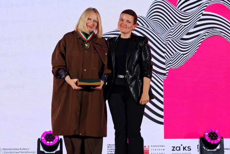 Odznaczona medalem Gloria Artis Katarzyna Nosowska oraz Ministra Kultury Marta Cienkowska stoją na scenie na tle biało-różowego ekranu.