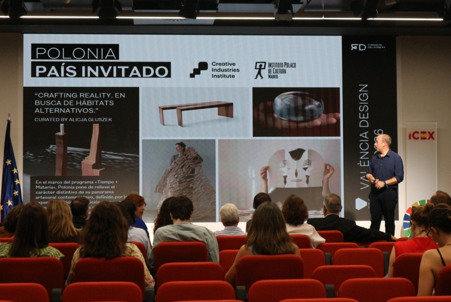 Ogłoszenie Polski, jako kraju gościnnego na Valencia Design Fest. Mężczyzna w granatowej koszuli przemawia prze kilkunastoosobową publicznością, a za nim jest prezentacja w języku hiszpańskim.