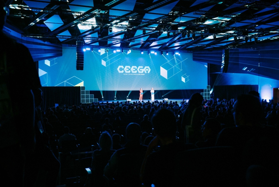 Szeroki kadru na scenę podczas gali CEEGA w dużej sali konferencyjnej. Na wielkim, podświetlonym na niebiesko ekranie widnieje logo CEEGA oraz geometryczne wzory kostek. Na środku oświetlonej scenie stoją dwie osoby prowadzące wydarzenie, a w przyciemnionej przestrzeni przed sceną widać sylwetki licznie zgromadzonej widowni.