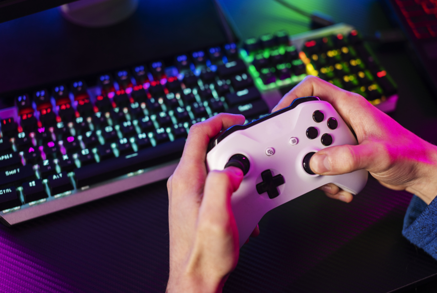 Ujęcie rąk gracza trzymających biały kontroler przed klawiaturą z podświetleniem RGB w dramatycznym różowo-fioletowym oświetleniu.