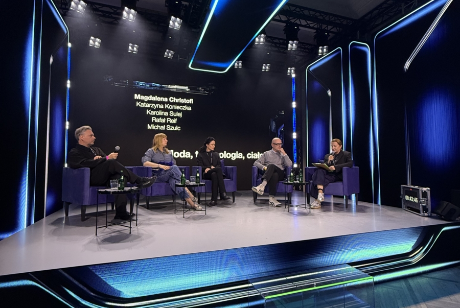 Pięć osób siedzi w fioletowych fotelach na nowoczesnej, oświetlonej na niebiesko scenie podczas panelu dyskusyjnego. W tle na dużym ekranie widać napis „moda, technologia, ciało” oraz listę prelegentów (Magdalena Christofi, Katarzyna Konieczka, Karolina Sulej, Rafał Reif, Michał Szulc). Osoba po prawej stronie prowadzi rozmowę, trzymając mikrofon i notatki.