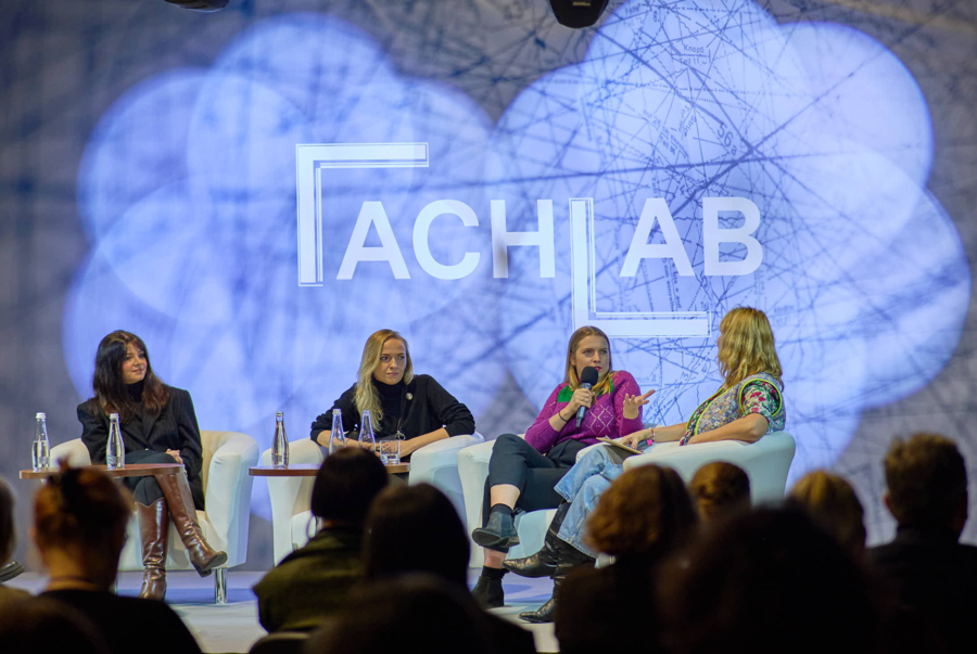 Cztery kobiety uczestniczą w panelu dyskusyjnym na scenie, siedząc w białych fotelach przed zgromadzoną publicznością. W tle na dużym ekranie widnieje biały napis „FACHLAB” na tle grafiki przypominającej wykroje krawieckie. Kobieta w różowym swetrze mówi do mikrofonu, gestykulując dłonią.