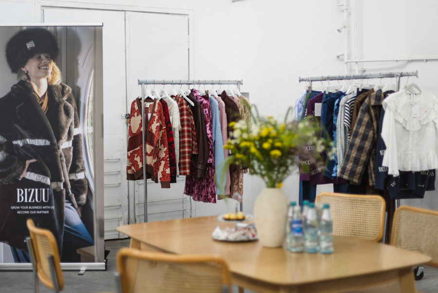 Jasny showroom marki BIZUU: na pierwszym planie drewniany stół z bukietem kwiatów, a w tle stojące wieszaki z kolorowymi ubraniami oraz baner reklamowy.