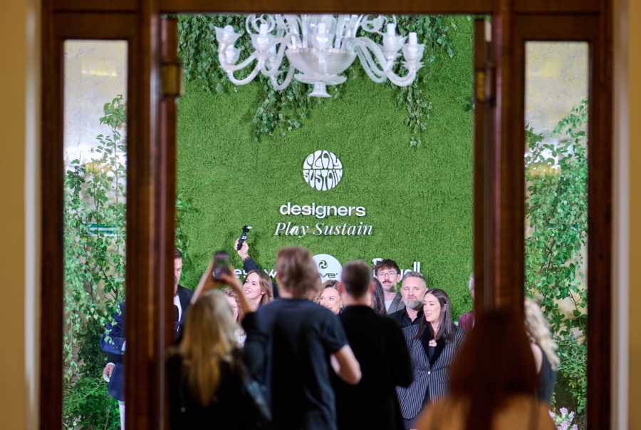 Widok przez otwarte drzwi na grupę osób pozujących na tle zielonej ścianki z napisem „Designers Play Sustain” i eleganckim szklanym żyrandolem.