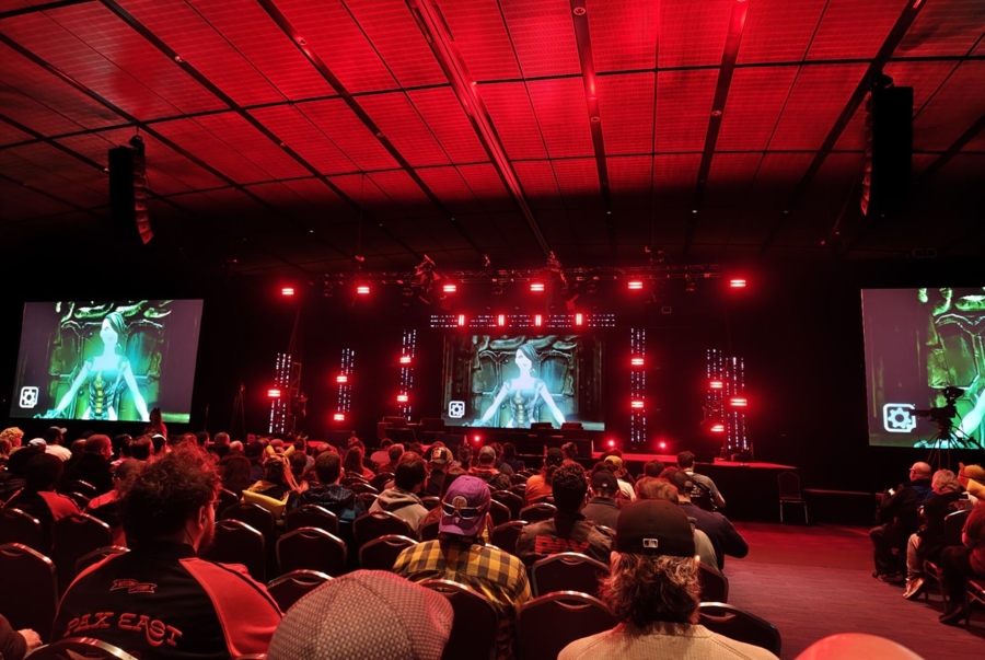 The audience sits in rows of chairs watching a panel on stage in a darkened conference room. The entire space is bathed in red lighting. Three large screens display a green-glowing female figure (possibly an android). One audience member in the foreground wears a jacket emblazoned with the words "PAX EAST."