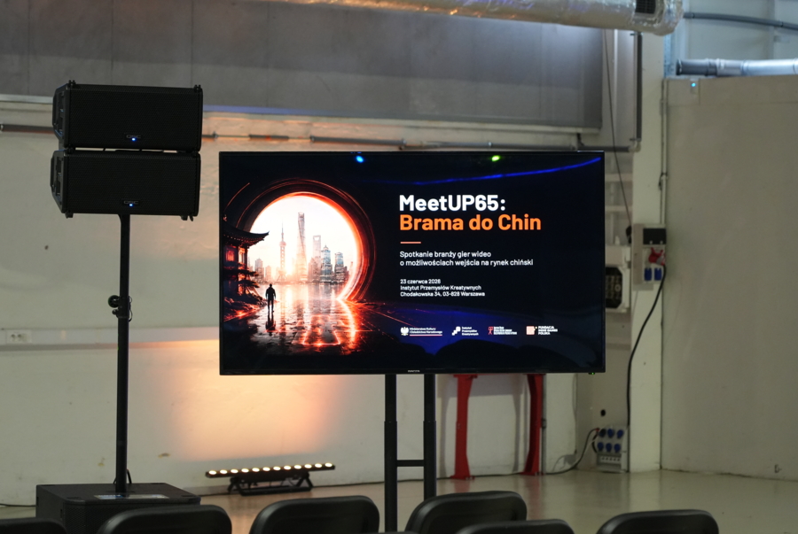 Duży ekran na statywie w sali konferencyjnej wyświetlający slajd wydarzenia „MeetUP65: Brama do Chin” z grafiką świetlistego portalu. Po lewej stronie stoi kolumna głośnikowa.
