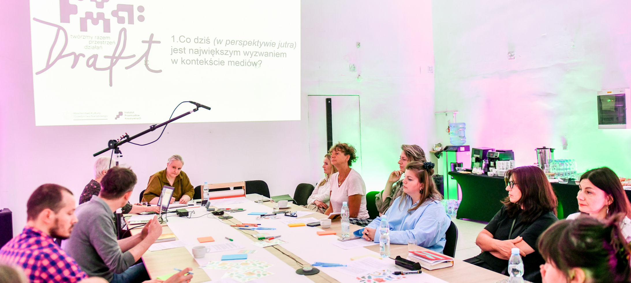 Uczestnicy warsztatów siedzący przy stole z materiałami do pisania i przysłuchujący się dyskusji. Na ścianie wyświetlany jest slajd prezentacji z pytaniem „Co dziś (w perspektywie jutra) jest największym wyzwaniem w kontekście mediów?”, a jasne wnętrze podświetlono różowym i zielonym światłem.