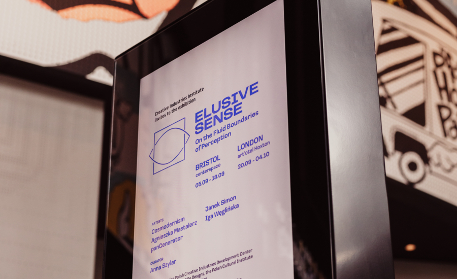 Zbliżenie na czarny pylon informacyjny z plakatem wystawy "ELUSIVE SENSE: On the Fluid Boundaries of Perception". Na białej planszy widnieją niebieskie napisy z terminami i lokalizacjami w Bristolu i Londynie (wrzesień-październik 20.09–04.10). Wymienieni są też artyści (Cosmodernism, Agnieszka Mastalerz, panGenerator, Janek Simon, Iga Węglińska) oraz kuratorka Anna Szylar. W tle rozmyte wnętrze z elementami komiksu.