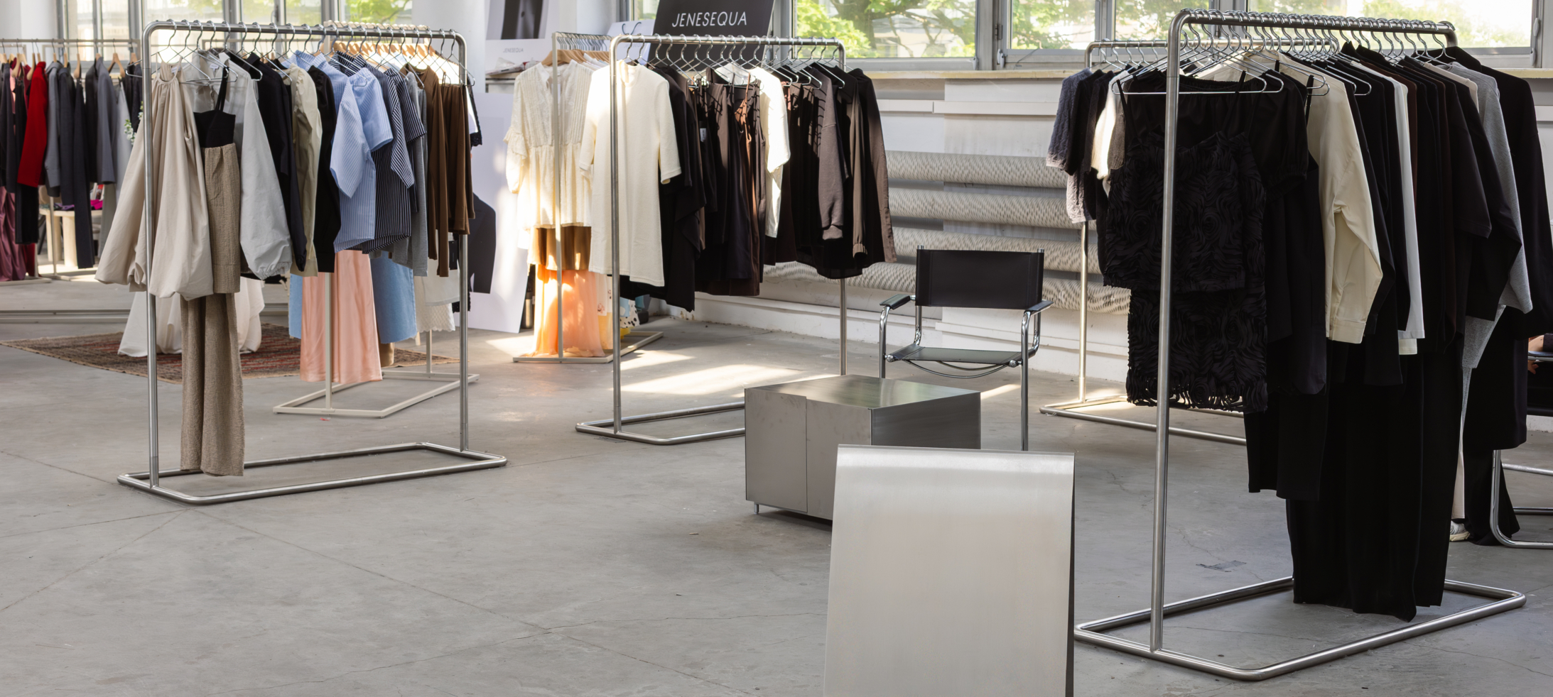 Minimalistyczny showroom modowy z metalowymi wieszakami pełnymi ubrań marek MUUV. i JENESEQUA w jasnej sali z dużymi oknami.