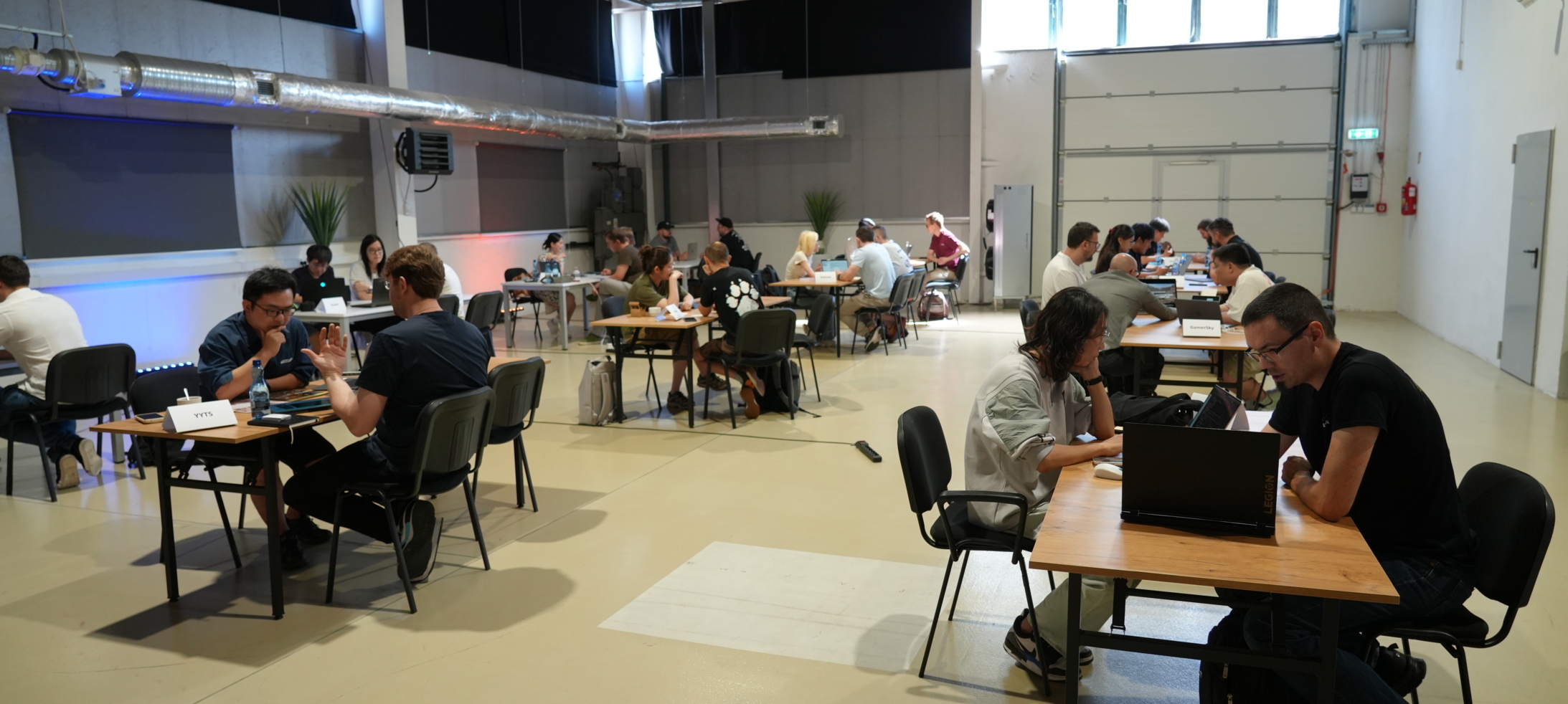 Przestronna, jasna hala w stylu przemysłowym z wieloma stolikami, przy których pary osób rozmawiają i pracują na laptopach podczas spotkań biznesowych.