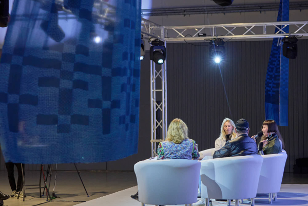 Cztery osoby uczestniczą w panelu dyskusyjnym, siedząc w białych fotelach na podwyższonej scenie. Z lewej strony zwisa duża, niebieska, półprzezroczysta tkanina we wzory, a nad sceną widoczne są metalowe kratownice z reflektorami.