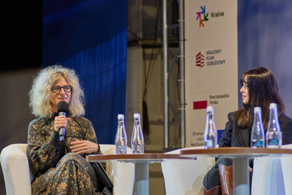 Dwie kobiety siedzą w białych fotelach na scenie podczas panelu dyskusyjnego. Kobieta po lewej w kręconych blond włosach mówi do mikrofonu, a po prawej przysłuchuje się jej kobieta w ciemnym żakiecie i brązowych kozakach. W tle widoczne są banery z logo FACHLAB oraz logotypami partnerów.