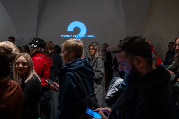 Biała ściana z niebieską dwójką i napisem Intencja czy odliczenie. Przed ścianą są ludzie, którzy na nią nie patrzą.