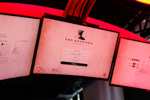 Trzy połączone monitory podświetlone czerwoną łuną. Na środkowym ekranie widoczne jest menu główne gry „Gra Szyfrów: Wojna o wszystko” z opcjami nawigacji, a na bocznych monitorach – schematy sterowania klawiaturą i myszą.