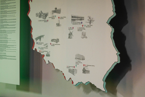 Duża, warstwowa rzeźba mapy Polski, zawieszona na czarnej kratownicy pod sufitu. Mapa jest biała z czerwonym konturem, na której umieszczono 15 ponumerowanych ilustracji kluczowych polskich zabytków i miejsc z opisami po polsku, m.in.: Żuraw (Gdańsk), Zamek Książąt Pomorskich (Szczecin), Rynek Wielki (Zamość), Wawel i Nowa Huta (Kraków), Dąb Bartek, Pomnik Syrenki (Warszawa). Po lewej widoczny jest pionowy panel z tekstem, a na dole fioletowa i niebieska pufa. Tło jest podświetlone i rzuca cienie.