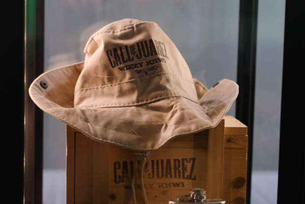 Beżowy materiałowy kapelusz z logo gry „Call of Juarez: Więzy Krwi” leżący na brandowanej drewnianej skrzyni wewnątrz szklanej gabloty. Przed skrzynią stoją gadżety z edycji kolekcjonerskiej: metalowa piersiówka oraz zapalniczka.