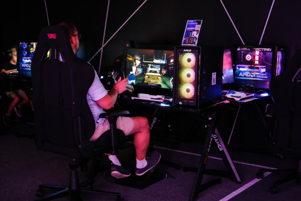 Mężczyzna siedzący na czarnym fotelu gamingowym Yumisu i grający w wyścigi samochodowe przy użyciu kierownicy z pedałami. Na biurku Pro-Gamer obok monitora stoi komputer z kolorowym podświetleniem RGB, a w tle widać sąsiednie stanowiska graczy w przyciemnionej sali.