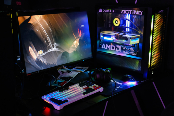 Stanowisko gamingowe z włączoną grą na monitorze i przeszkloną obudową komputera Actina z podświetleniem RGB. Na biurku znajdują się jasna, podświetlana klawiatura mechaniczna, czarne słuchawki wokółuszne oraz myszka z wielokolorowym podświetleniem.