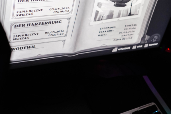 Monitor wyświetlający menu zapisów gry w formie otwartej książki z tytułem „Der Harzerburg” i ilustracją sterowca. W dolnej części zdjęcia widoczna jest klawiatura mechaniczna z zielonym podświetleniem LED w ciemnym otoczeniu.