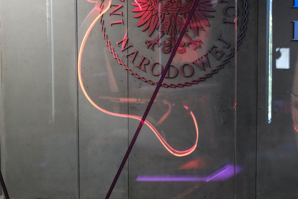 Logo Instytutu Pamięci Narodowej z polskim orłem na szarej ścianie, widoczne przez szklaną ściankę z czerwonymi refleksami świetlnymi. Na pierwszym planie nieostry fragment czerwonego biurka z klawiaturą.