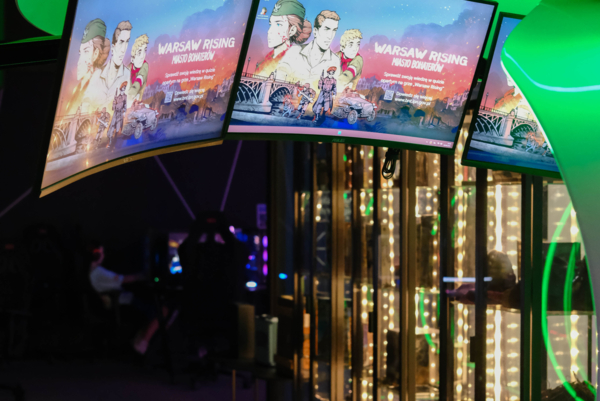 Trzy monitory w futurystycznej kapsule gamingowej z zielonym podświetleniem LED, wyświetlające grafikę gry „Warsaw Rising: Miasto Bohaterów”. Na stanowisku widoczne jest biurko z klawiaturą i myszką, a w tle podświetlone gabloty ekspozycyjne.
