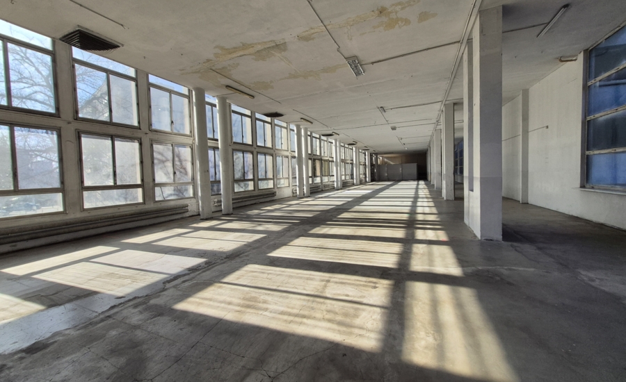 Długa, pusta sala industrialna z rzędem okien i słońcem wpadającym na betonową podłogę.