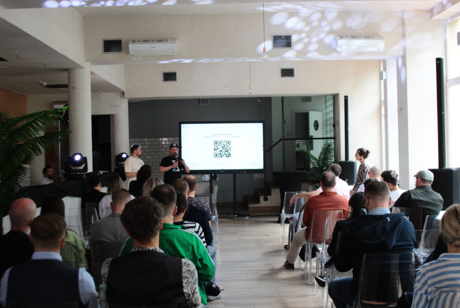 A meeting in a bright room. A man with a microphone is addressing an audience seated on chairs, while a slide displaying a QR code appears on the screen behind him.